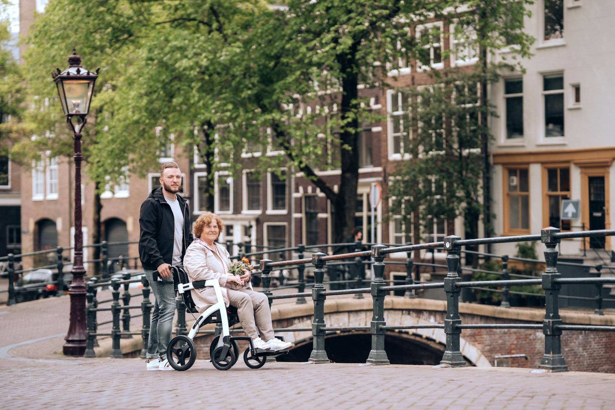 Senior woman using a rollator on a canal bridge in Amsterdam with a young man assisting. Urban scene.